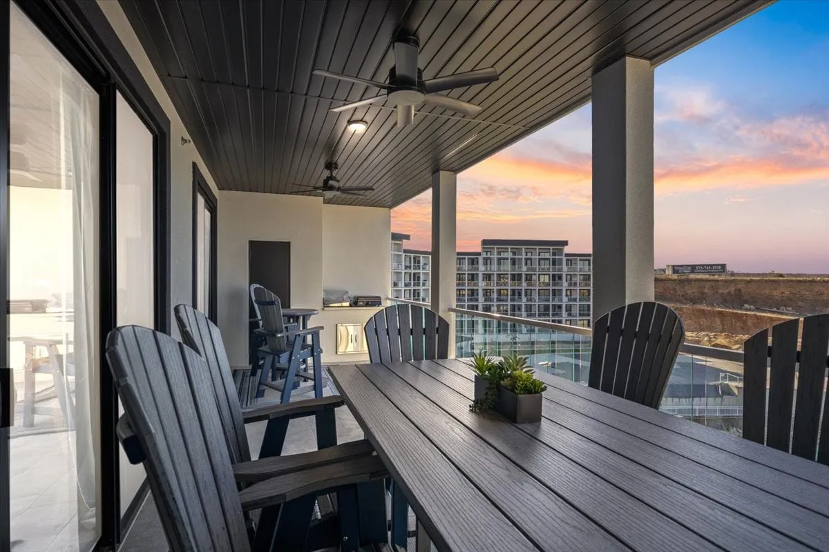 This exterior image features a spacious balcony set up for outdoor dining, complete with an elegant table, comfortable chairs, and overhead ceiling fans. The balcony overlooks a stunning scenic lake view, inviting relaxation and social gatherings as the sun sets over the water.