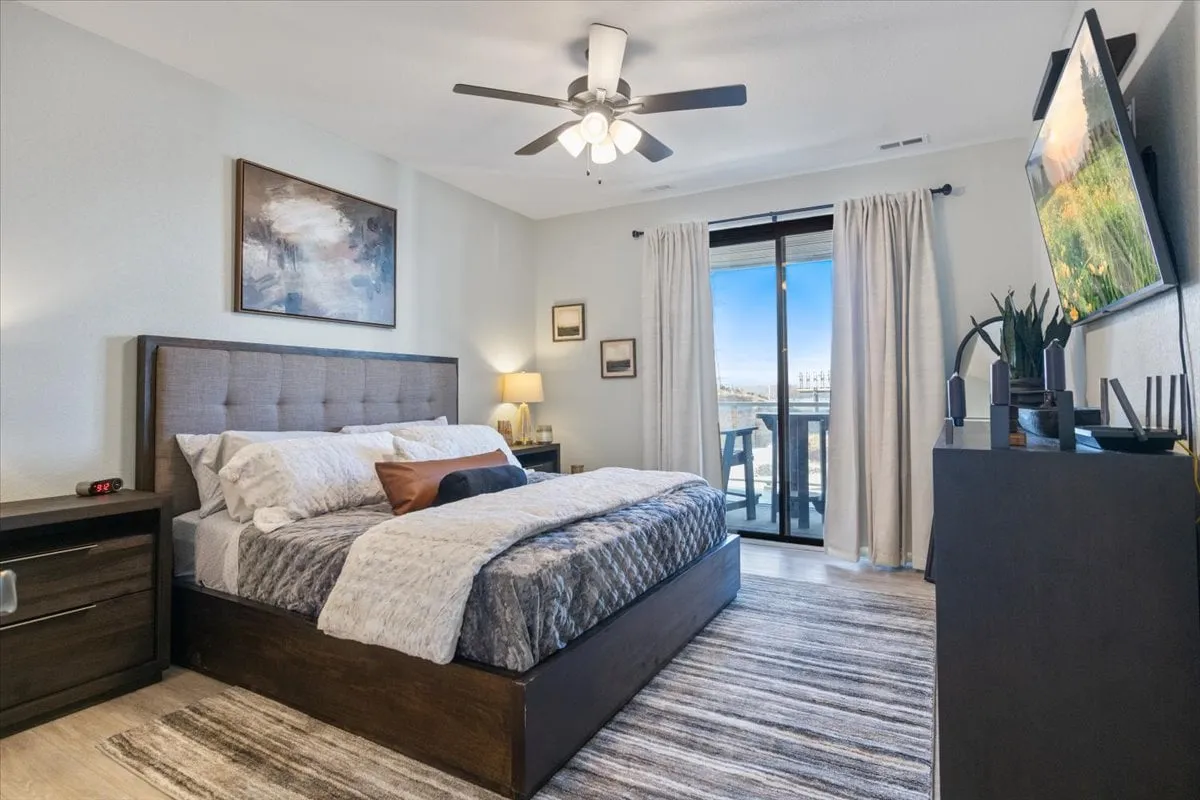 A calming bedroom scene featuring a neatly made bed with plush bedding, a balcony door letting in natural light, a television, and tasteful artwork above the nightstand. The room feels restful and inviting, and the balcony door hints at an exterior view—possibly the lake.