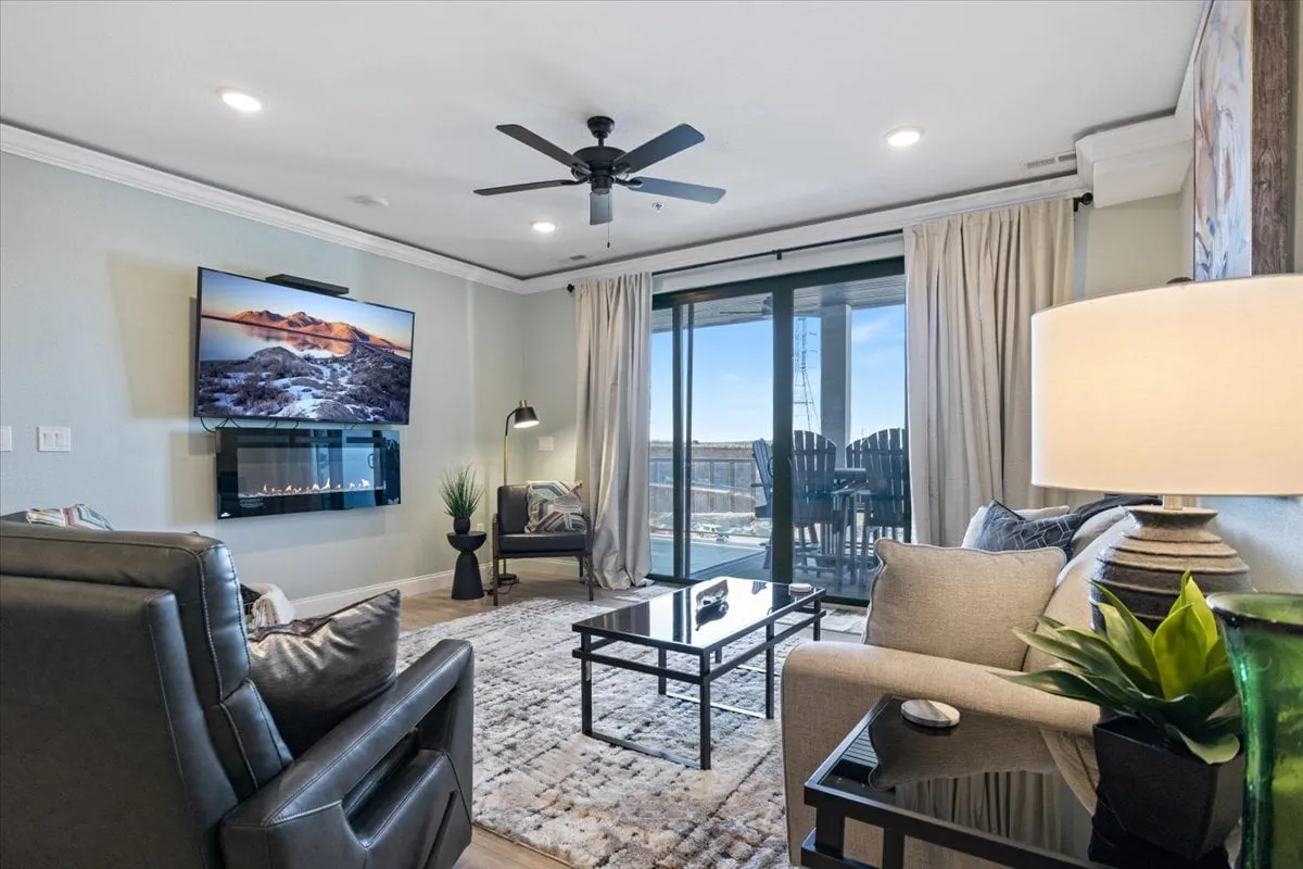 A cozy living area with comfortable seating faces a large TV showing a scenic landscape, while a modern fireplace and broad glass doors lead directly to the balcony. Decorative plants add a touch of homeliness, and the view through the doors hints at the water beyond, tying the interior’s tranquility to the outdoor beauty.