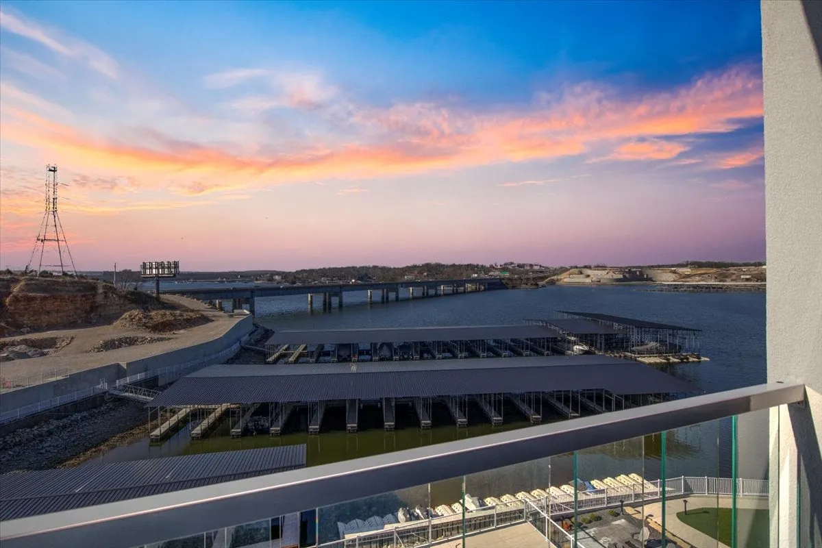 A panoramic lakefront view at sunset, with marina docks, a distant bridge, and tranquil waters. Boaters and people relaxing by the water can be seen, and the sky is painted in vibrant sunset hues.