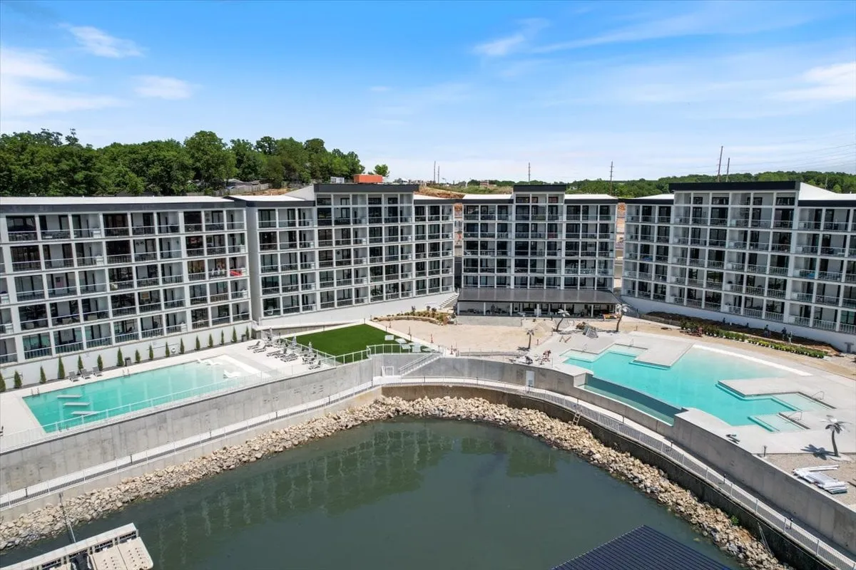 A wide view of modern condo buildings set against sparkling swimming pools, lush lawns, and a waterfront area. The inviting outdoor spaces suggest summer fun and community events, with ample room for sunbathing or social activities by the water.