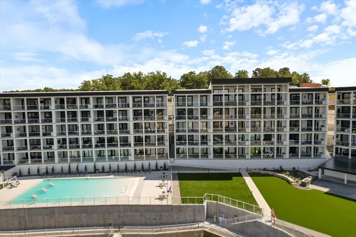 A lively communal pool area surrounded by balconies, green lawn, and the building's modern exterior. The pool is perfect for socializing, sunbathing, or enjoying a quiet morning, with the lake just beyond.