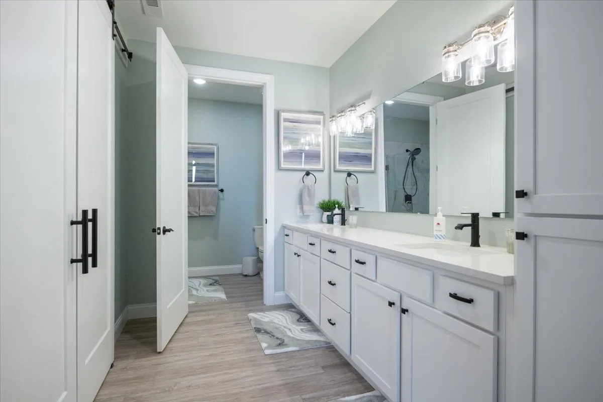 A modern bathroom with a double vanity, elegant mirrors, contemporary light fixtures, and decorative art accents. The space appears organized, serene, and offers ample room for personal care routines.