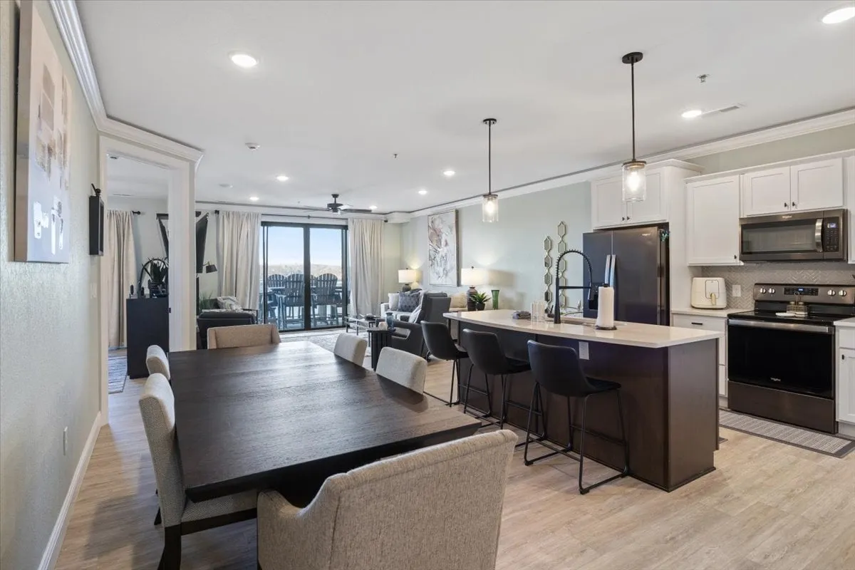 This interior shot captures a spacious open-plan area featuring a dining table, kitchen island, and living area, all illuminated by large windows that reveal shimmering water views beyond. The scene suggests a welcoming space that accommodates gatherings, shared meals, and relaxation, while the abundant natural light and visual access to the outdoors reinforce the panoramic connection to the lake.