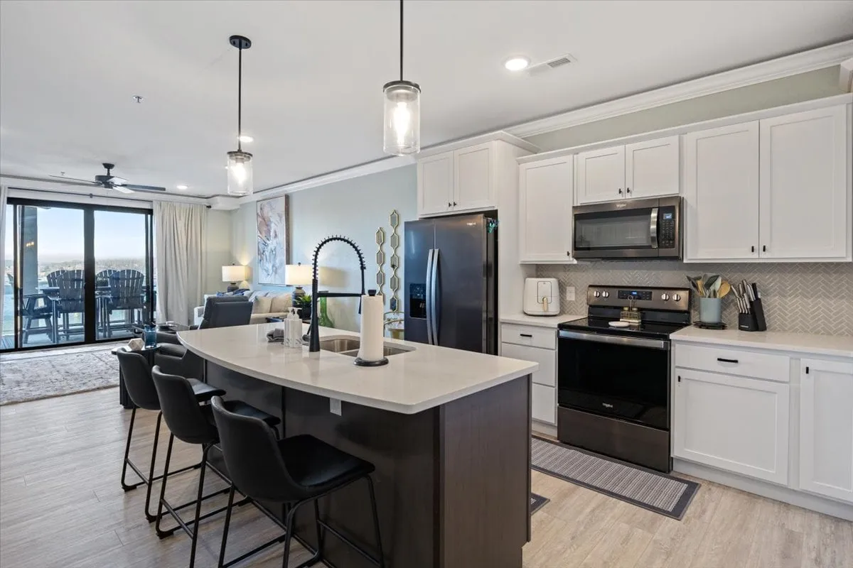 A modern, spacious kitchen with a central island, black appliances, large windows letting in plenty of natural light, and bar stools lined up for socializing. The space feels open and well-equipped, ideal for cooking, gathering, and enjoying a morning coffee or evening meal.