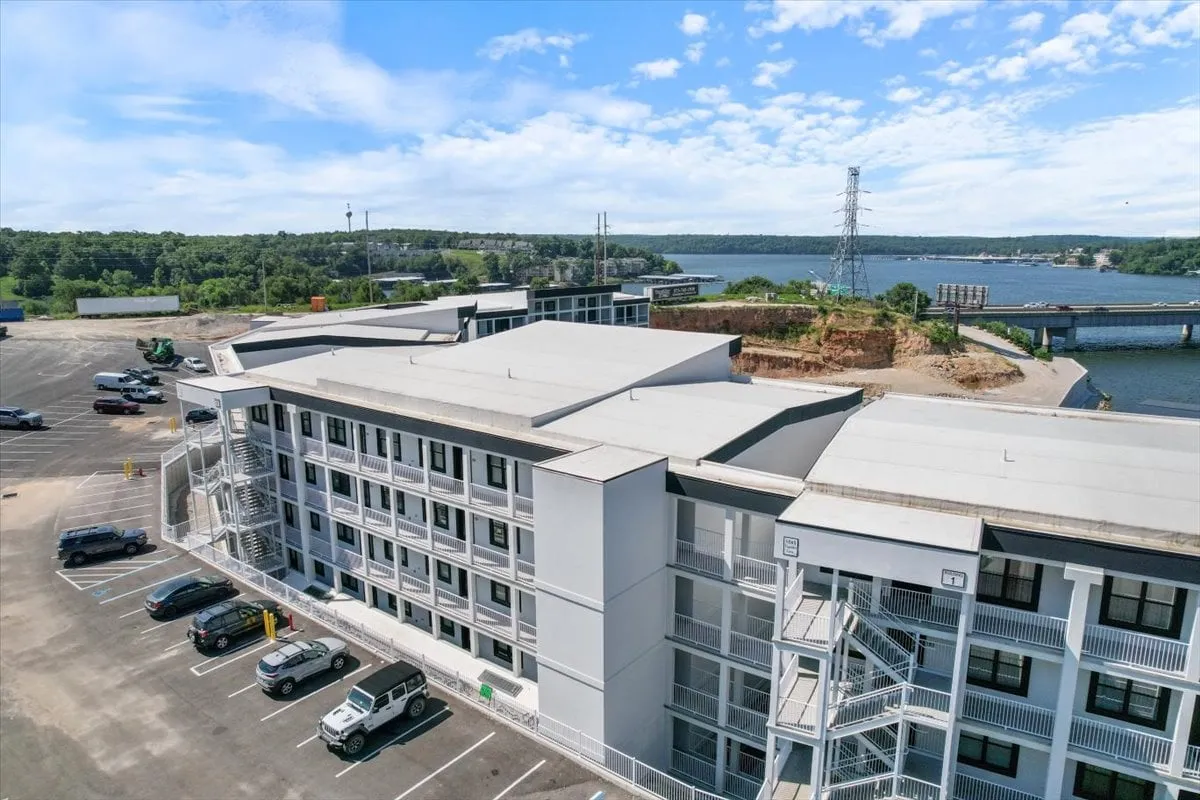 A wide exterior view of the property, with the building structure and parking area in the foreground and the river shimmering in the background. Lush greenery surrounds the property, offering a sense of both convenience and natural beauty.